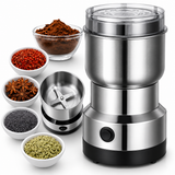 A stainless steel electric spice grinder with a clear lid and black base, shown alongside bowls of ingredients including ground brown spice, red pepper flakes, star anise, black seeds, and green fennel seeds on a white background.